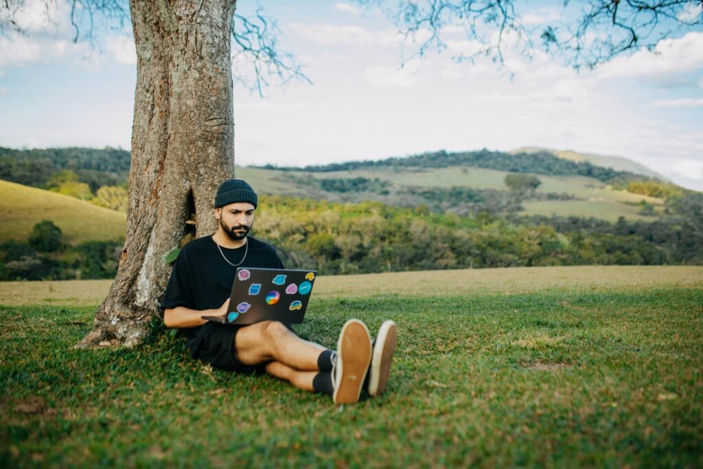 A young man sitting under a tree, working on a laptop in a serene meadow setting.